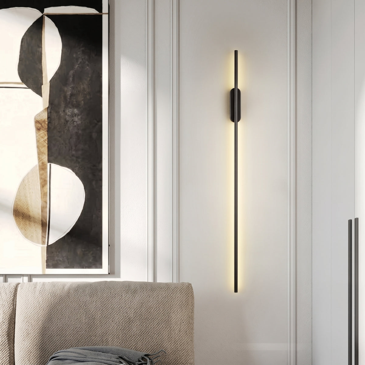 A slender black vertical wall lamp glowing soft light on a wall beside a large black-and-white abstract painting and a beige sofa.