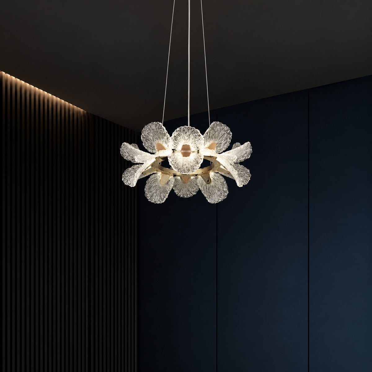A modern flower-shaped pendant light with textured clear glass petals and a brushed gold center, suspended in a dark interior with navy paneled walls and a slatted accent wall.