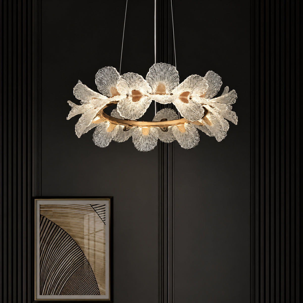 A modern floral chandelier with textured white glass flower petals and a gold-toned circular frame, suspended against a dark paneled wall with a framed abstract print below.